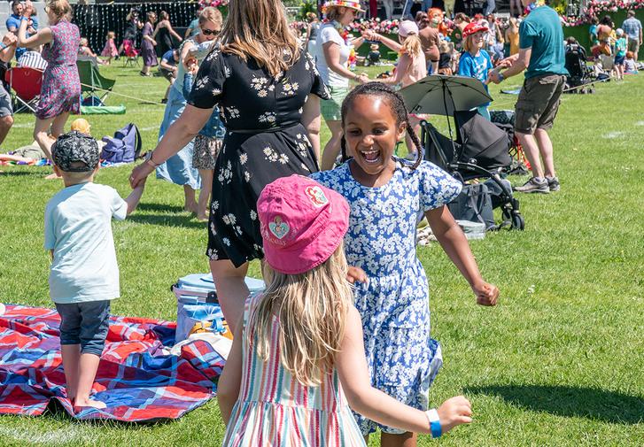 Children and grown-ups dance around on Oaklands Park. In the middle of an image a girl with pigtails smiles and dances with a girl in a pink hat.