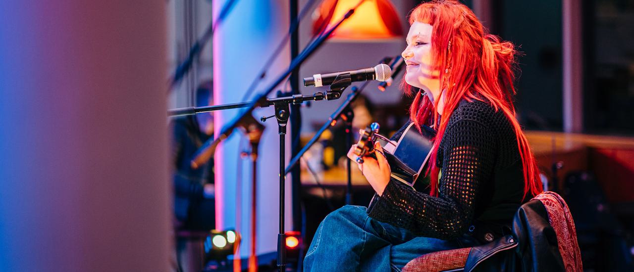A young woman with long, bright red hair sits at a microphone holding a guitar. She wears a black top with lattice sleeves and jeans, and faces the left, smiling, with her face illuminated by a warm spotlight.