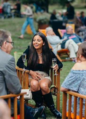 A group of people sit on colourful deckchairs in the park, chatting and smiling.