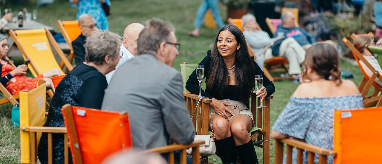 A group of people sit on colourful deckchairs in the park, chatting and smiling.