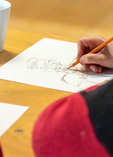 A woman sketches a pencil drawing of a man onto a sheet of paper. The paper is on a wooden table, and the pencil is orange.