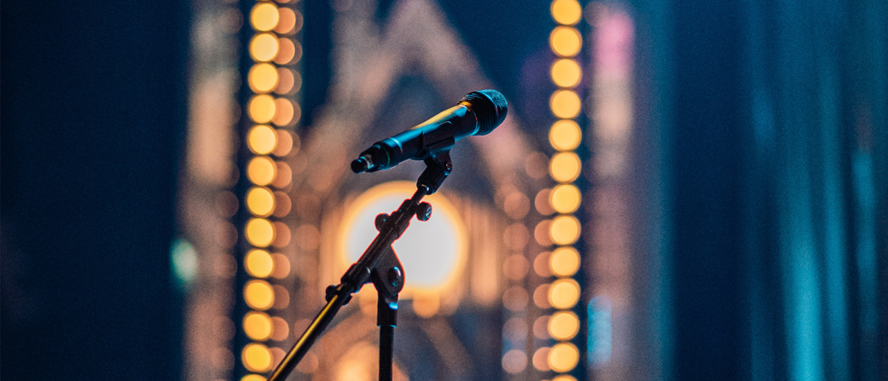 A microphone is in the centre of the frame, in front of a backdrop of blurred golden lights.