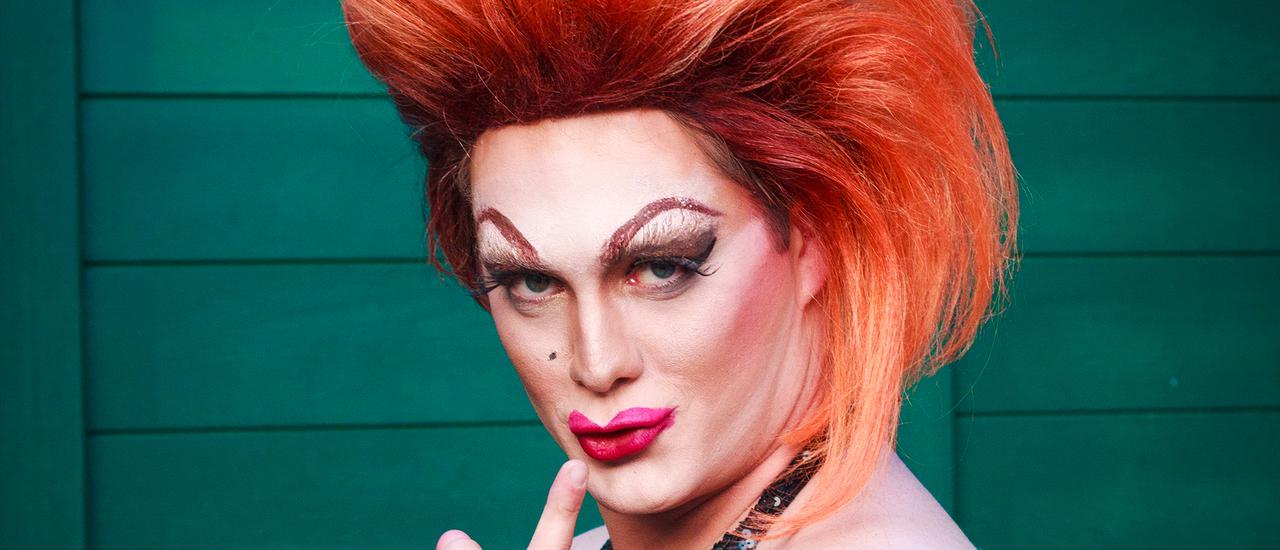 A drag queen wearing a large, permed red wig stands in front of a green wall. She has drawn-on exageratted eye brows, and pink lipstick. She holds a finger in front of her mouth and purses her lips, 'shh'. She is wearing a dark blue velvet shawl.