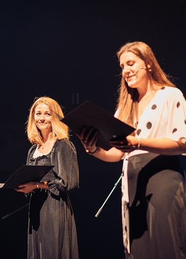 A smiling woman with long brown hair stands on stage, reading from a large black book.. She wears a white top with black spots, and black trousers. She is wearing a microphone on her face. To the left, a blonde woman is looking towards her, smiling. This woman is in a long black dress and holds another book.