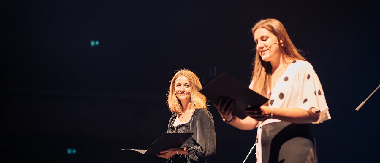 A smiling woman with long brown hair stands on stage, reading from a large black book.. She wears a white top with black spots, and black trousers. She is wearing a microphone on her face. To the left, a blonde woman is looking towards her, smiling. This woman is in a long black dress and holds another book.