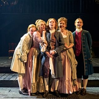 Five young actors stand huddled together on stage, looking up in front of them with excited expressions. The actor in the centre is wearing a long dark jacket, white socks and black shoes with buckles. Either side of them two actors in dresses lean in affectionately. On the left a shorter actor in a white jacket rests their head on the shoulder of another. On the right stands someone in a long blue jacket, slightly seperated from the group.