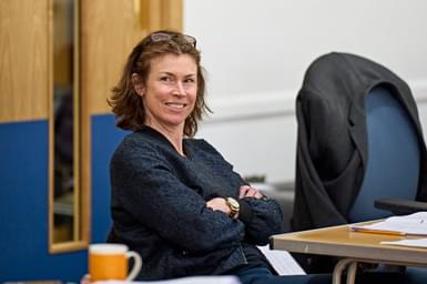 A brown-haired woman sits at a table, smiling at something out of frame. She has glasses on her head, is wearing a gold watch and has her arms crossed. She is wearing a black jumper. On the table in front of her is a script, a pencil and a mug of tea.