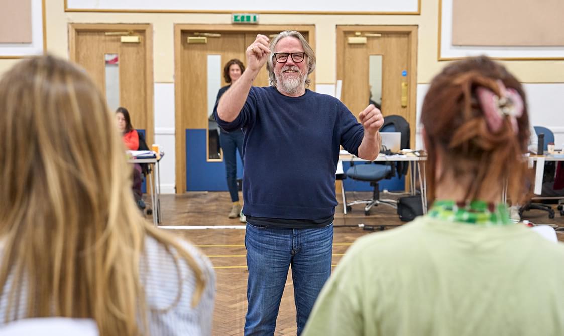 A man with a grey beard and glasses stands in a rehearsal room, smiling and holding his arms up in front of him. He is gesticulating like he is conducting an orchestra. In the foreground are the backs of two people's heads, as they look towards the man.