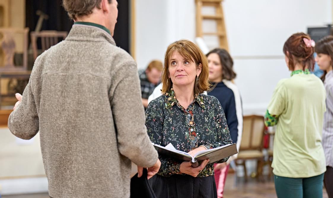 A woman in a floral shirt stands in a rehearsal room, holding a large book open in frotn of her. She has shoulder-length brown hair and is looking up at a man in front of her, who has his back to the camera. He is wearing a brown jumper, and in the background other people are stood around, talking.