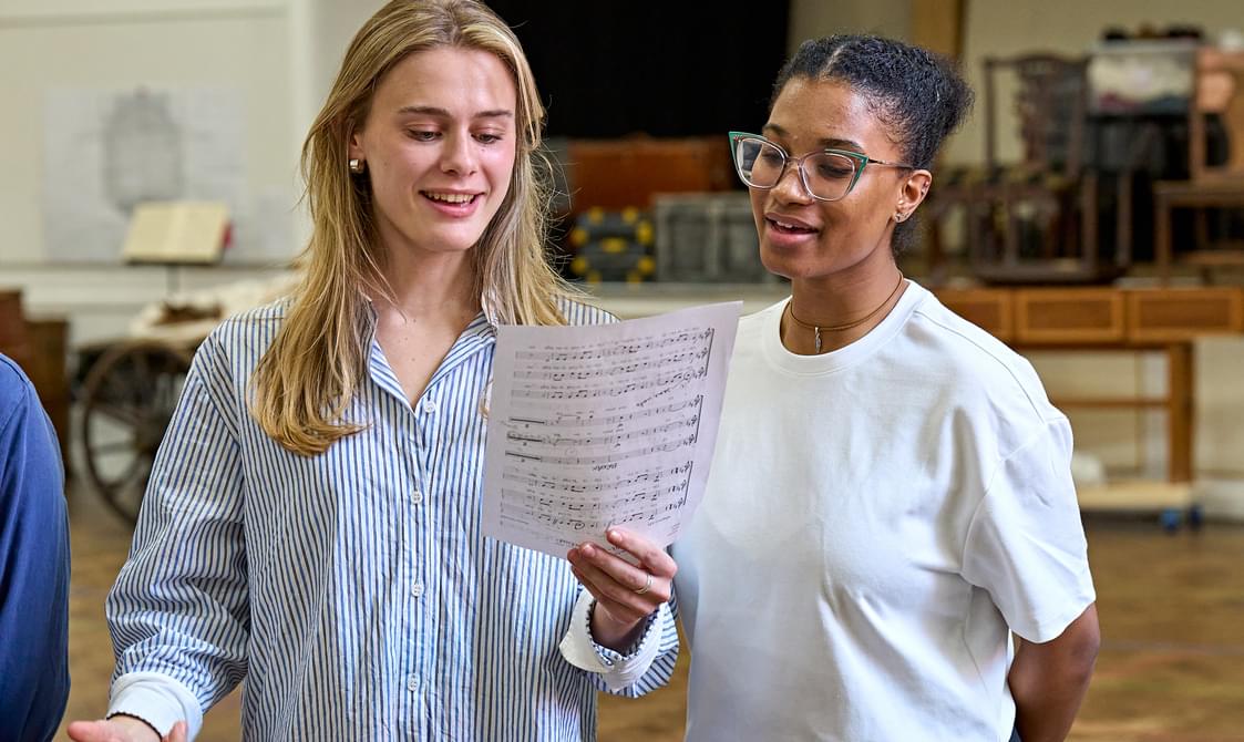 A young woman wearing a blue and white striped shirt, with long blonde hair, stands in a rehearsal room. She is holding a page of sheet music in front of her and is looking at it, smiling and signing. On the right is another young woman, also looking at it and singing. She has green-rimmed glasses and wears a white t-shirt.