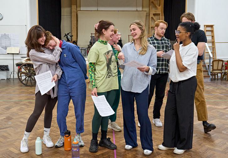 A group of actors are stood together, smiling. On the left, one has their head on another's shoulder, in the middle three people are laughing together. In the background, two actors look at a script. On the floor are three water bottles.