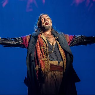 A man stands alone with his arms spread wide open in song. He has long black hair and a thick black beard. He wears a layered collection of well-worn clothes.