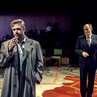 Two men stand on a stage that looks like a war map. The man in the foreground wears a long trench coat and smokes a cigarette. The man in the midground wears a smart suit and striped tie.