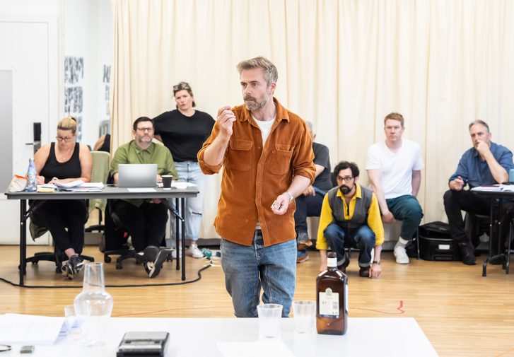 A man in an orange overshirt and jeans stands behind a table, his right arm gesturing upwards. Behind him, a number of other people sit at tables or crouching on the floor, looking on.