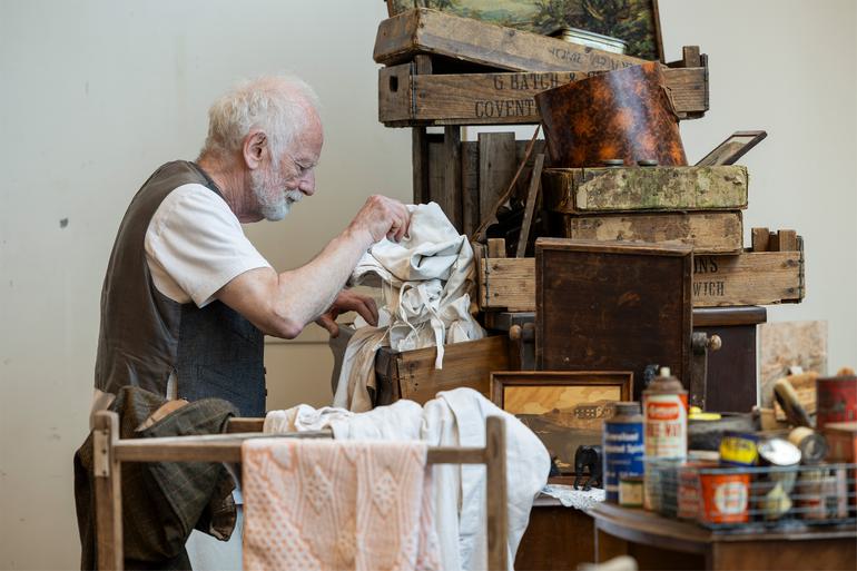 Actor Ian McDiarmid stands next to a tall pile of wooden crates, paintings and boxes in a rehearsal room. He is looking through a messy pile of white clothes or bedding. In front of him is a clothes drying rack and a table covered in paint and spray cans.