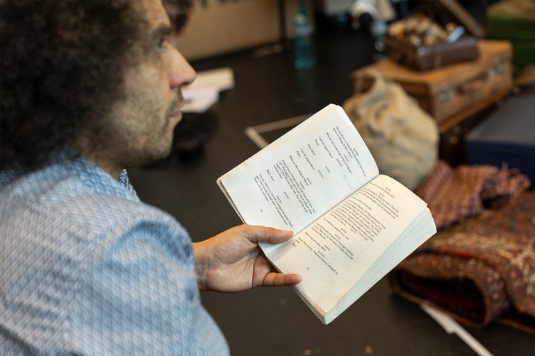 Director Justin Audibert holds a playscript in his hand, and looks out at a rehearsal room. Blurry in the background, piled up rugs, boxes and bags can be seen.