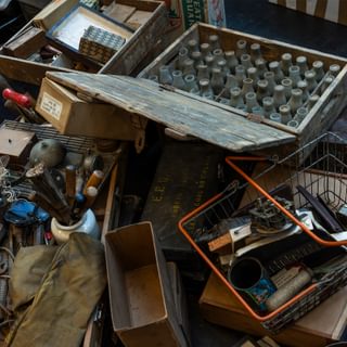 Cluttered boxes cover the floor in a rehearsal room, with the camera looking down on them. Including: a scuffed wooden toolbox filled with rusty pliers, hammers and chisels; a crate filled with dirty glass bottles; a shopping basket with paint cans and rollers.