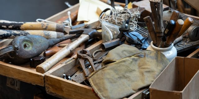 A scuffed wooden box is filled with metal tools: Rusty pliers, files, a large hammer, a pot of chisels and screwdrivers, a ball of string.