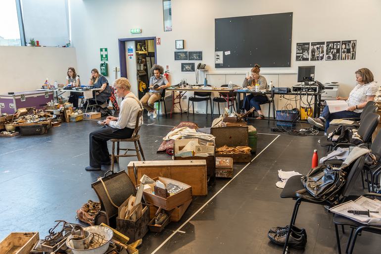 A rehearsal room, with desks and chairs around the edge of the room and piles of clutter across the floor. There are rugs, boxes, piles of paper, scraps of wood and metal. An actors sits on a chair in the centre of the room, facing away from the camera.