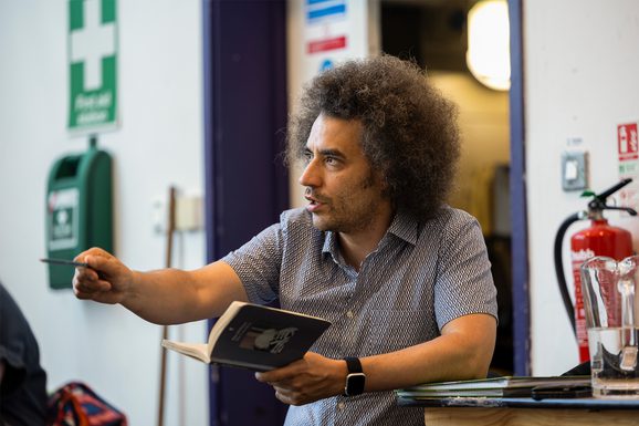 Director Justin Audibert leans his elbow on a table in a rehearsal room. He holds a book in his hand and points with a pencil in his other hand. He has an engaged look on his face and is talking. He is wearing a blue patterned shirt.