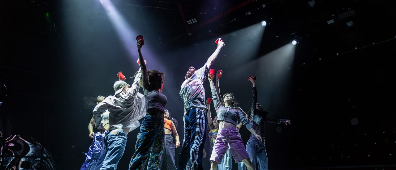 A group of young people dance on top of a kitchen island on a stage. They are all jumping in unison, with their feet off the ground and an arm stretched above their head holding a plastic cup. They are wearing colourful, sporty clothes - trainers, jogging bottoms and shorts. They are lit with dramatic white spot lights.