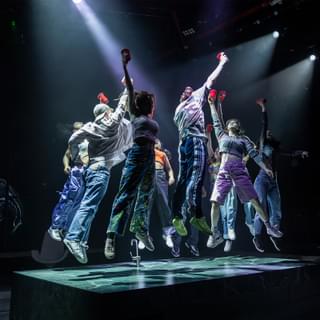 A group of young people dance on top of a kitchen island on a stage. They are all jumping in unison, with their feet off the ground and an arm stretched above their head holding a plastic cup. They are wearing colourful, sporty clothes - trainers, jogging bottoms and shorts. They are lit with dramatic white spot lights.