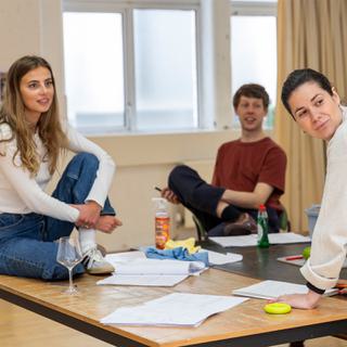Three people are in a rehearsal room, all smiling at something out of frame. On the left is a woman in jeans and a white top sat cross legged on a table, in the centre is a man in a red t-shirt sat on a chair, and on the right is a woman in a white jumper leaning on the table. On the table are paper scripts and rags and cleaning bottles that you would find in a kitchen.