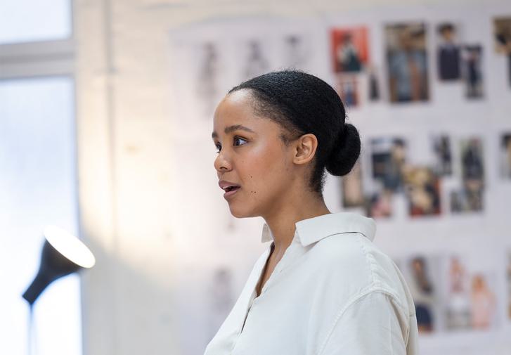A young woman in a white shirt and black hair in a bun stands in a rehearsal room. She is holding a black notebook, with a shocked look on her face, her mouth open slightly as she looks off camera.