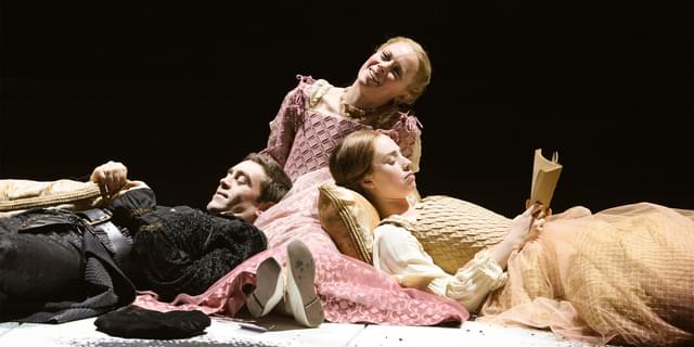 One man and two women dressed in traditional Tudor costume lie down and recline on cushions. One of the women is reading and they all seem comfortable and happy in each other's company