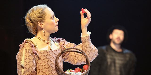 An actress in traditional Tudor costume picks a strawberry out of a basket and holds it up. A male actor in traditional Tudor costume stands in the background and is blurred.