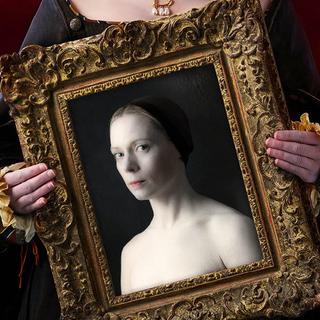 A woman's hands can be seen holding an ornate golden picture frame. The bottom of the woman's face is visible above the frame, she has a slight smile, red lipstick and is wearing a pearl neckace with a golden 'B' on it. Inside the picture frame is an image of a serious looking woman. The top of her chest and shoulders are exposed, and she wears a brown cap with her hair pulled back.