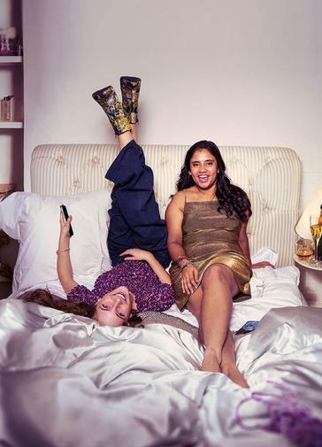 Two teenage girls sit on an rumpled bed with a pale pink satin duvet, smiling at the camera. The girl on the left lies on her back, with her feet up against the headboard, holding a phone in one hand. She wears patterned high heel noots, blue trousers and a purple ruffled top. To the right is a girl in a golden dress, leaning against the headboard with one foot crossed over the other.