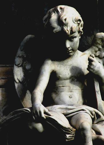 A statue of a curly haired cherub, holding some papers in his hand. The statue is in shadows, with the left side of the face and half its wing in darkness. It looks dramatic.