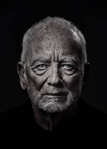 An elderly man (Ian McDiarmid) stares at the camera. His face is wrinkled and looks stern and dramatic, with shadows under his eyes and chin. He is wearing a black shirt, and his face in lit in a silvery colour.