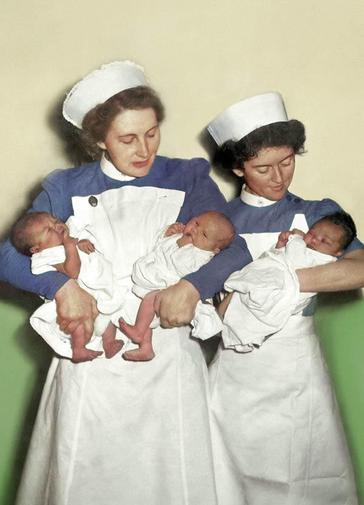 Two midwives in white smocks, white caps and blue blouses stand against a green and white wall. The midwife on the left holds two newborn babies, they look like twins. The midwife on the right holds one newborn baby with dark hair. They both look down caringly at the babies.