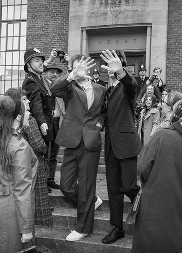 A black-and-white-photo taken on the crowded steps of Chichester Law Court. In the centre, two young men in stylish suits walk down the steps, holding their hands in front of their faces to block the camera. Around them, women in 1960s hairstyles and coats look up at the two men, some with excited expressions. On the left stands a stern policeman in a hat, with two more policemen and photographers in the background.