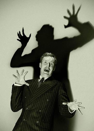 A black and white image of a man in a pin stripe suit looks aghast with his arms and hands outstretched. Behind him his shadow lurks overhead ready to pounce.