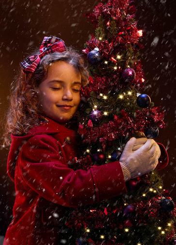 A young girl in a red winter coat hugs a small Christmas tree. She is smiling contentedly with her eyes closed, and has a red tartan bow in her hair. The tree is decorated with red tinsel, purple baubles and gold twinkly lights. Snow is falling around them.