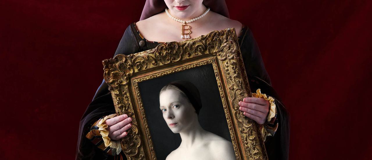 A woman's hands can be seen holding an ornate golden picture frame. The bottom of the woman's face is visible above the frame, she has a slight smile, red lipstick and is wearing a pearl neckace with a golden 'B' on it. Inside the picture frame is an image of a serious looking woman. The top of her chest and shoulders are exposed, and she wears a brown cap with her hair pulled back.