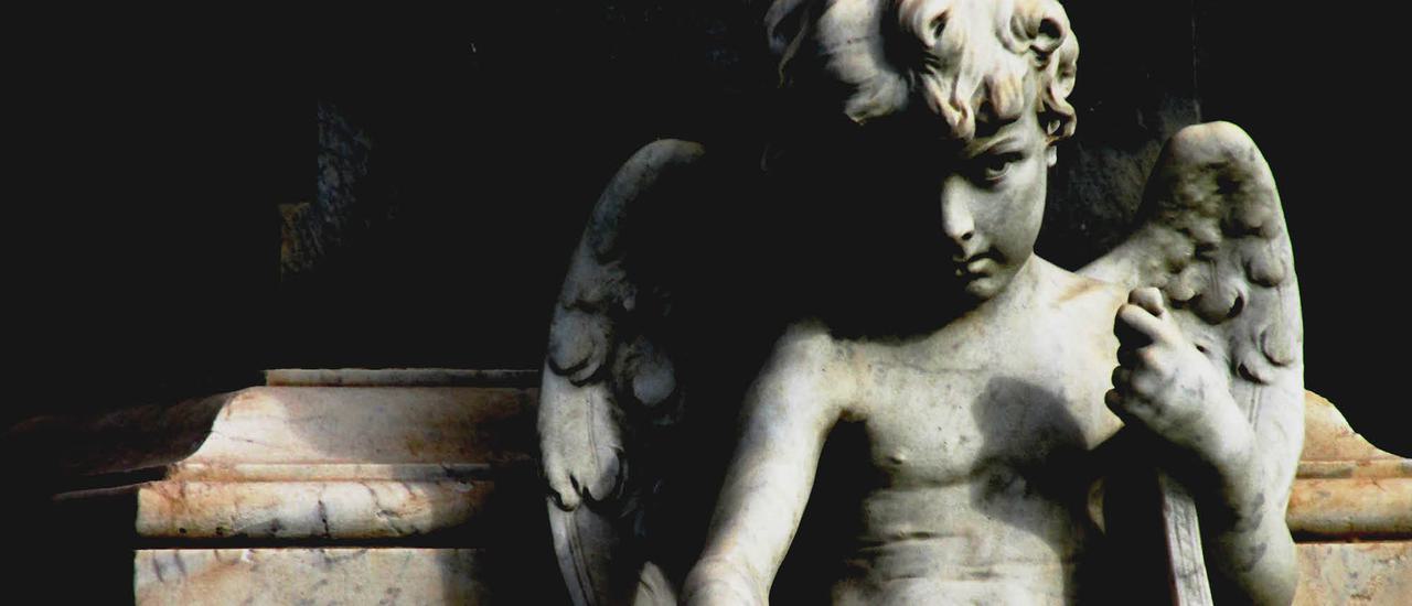 A statue of a curly haired cherub, holding some papers in his hand. The statue is in shadows, with the left side of the face and half its wing in darkness. It looks dramatic.