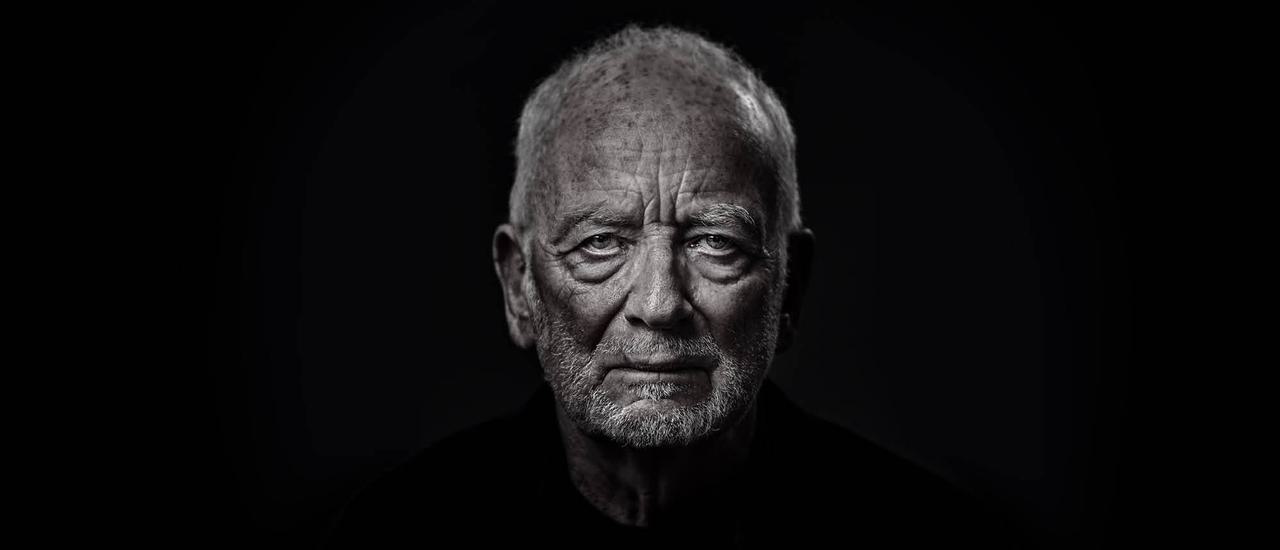 An elderly man (Ian McDiarmid) stares at the camera. His face is wrinkled and looks stern and dramatic, with shadows under his eyes and chin.  He is wearing a black shirt, and his face in lit in a silvery colour.
