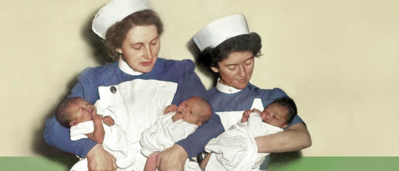 Two midwives in white smocks, white caps and blue blouses stand against a green and white wall. The midwife on the left holds two newborn babies, they look like twins. The midwife on the right holds one newborn baby with dark hair. They both look down caringly at the babies.