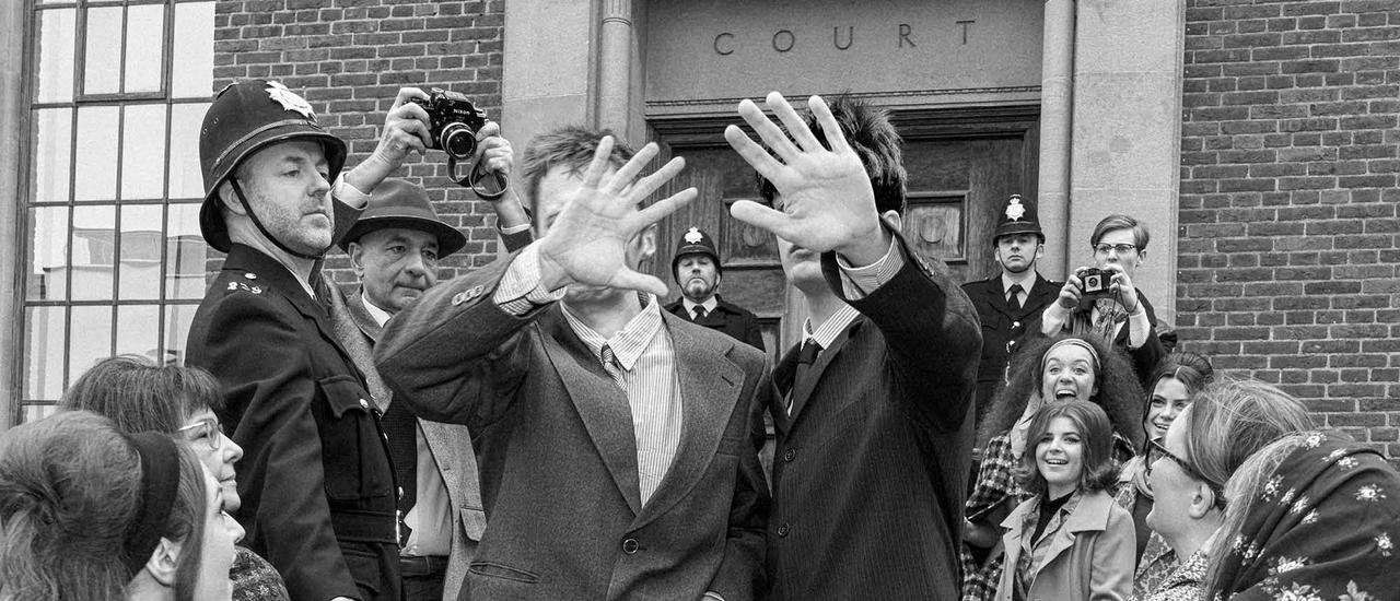 A black-and-white-photo taken on the crowded steps of Chichester Law Court. In the centre, two young men in stylish suits walk down the steps, holding their hands in front of their faces to block the camera. Around them, women in 1960s hairstyles and coats look up at the two men, some with excited expressions. On the left stands a stern policeman in a hat, with two more policemen and photographers in the background.