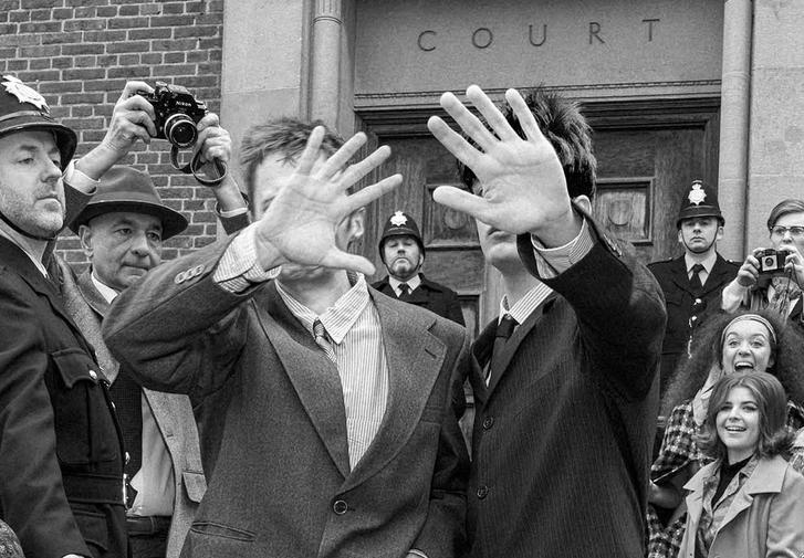 A black-and-white-photo taken on the crowded steps of Chichester Law Court. In the centre, two young men in stylish suits walk down the steps, holding their hands in front of their faces to block the camera. Around them, women in 1960s hairstyles and coats look up at the two men, some with excited expressions. On the left stands a stern policeman in a hat, with two more policemen and photographers in the background.