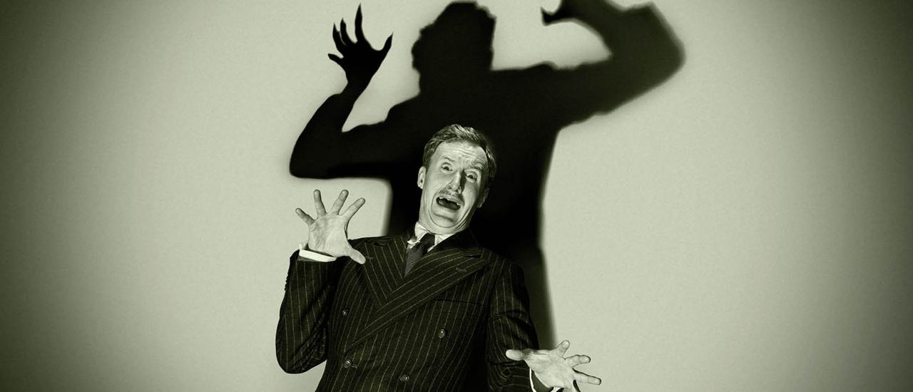 A black and white image of a man in a pin stripe suit looks aghast with his arms and hands outstretched. Behind him his shadow lurks overhead ready to pounce.