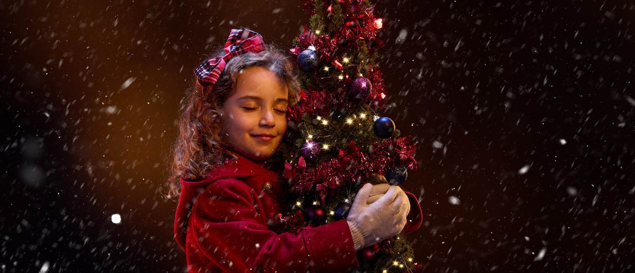 A young girl in a red winter coat hugs a small Christmas tree. She is smiling contentedly with her eyes closed, and has a red tartan bow in her hair. The tree is decorated with red tinsel, purple baubles and gold twinkly lights. Snow is falling around them.