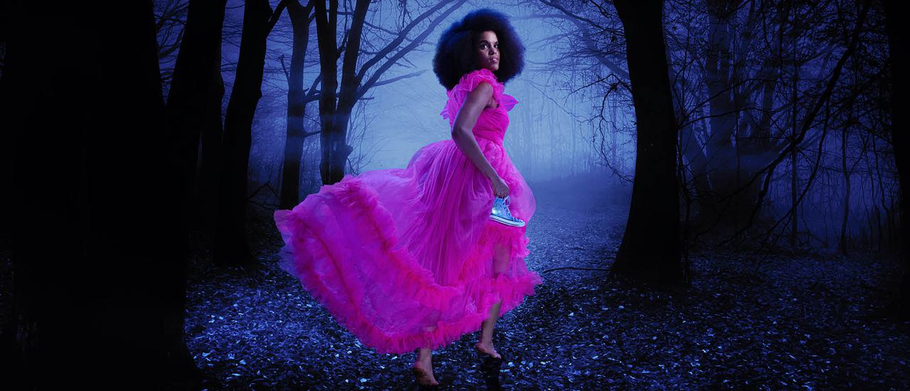 A woman in along, ruffled pink chiffon dress runs through a dark, misty woodland away from the camera. She is turning to look behind her, has an afro framing her face, and is barefoot. She holds a silver trainer in her hand. She is surrounded by mysterious looking trees and branches.