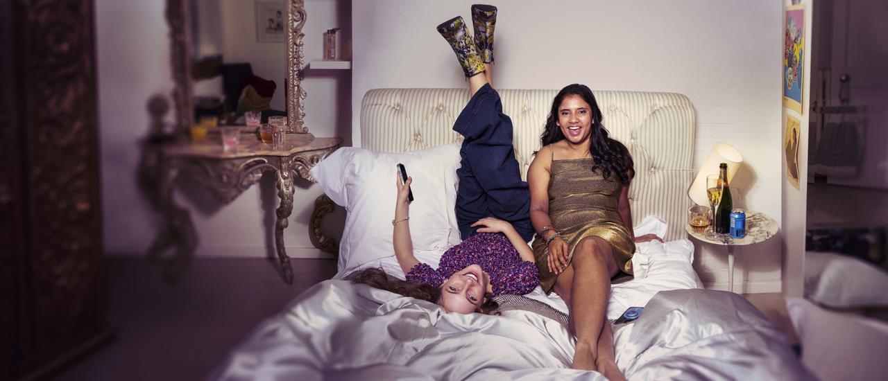 Two teenage girls sit on an rumpled bed with a pale pink satin duvet, smiling at the camera. The girl on the left lies on her back, with her feet up against the headboard, holding a phone in one hand. She wears patterned high heel noots, blue trousers and a purple ruffled top. To the right is a girl in a golden dress, leaning against the headboard with one foot crossed over the other.