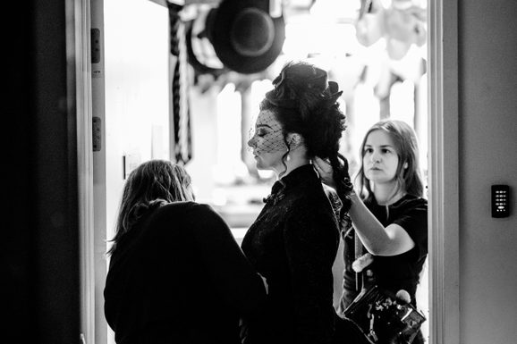 A performer stands side-on, eyes closed as though preparing calmly to go on stage. Two wardrobe assistants perfect the performer's wig and costume.