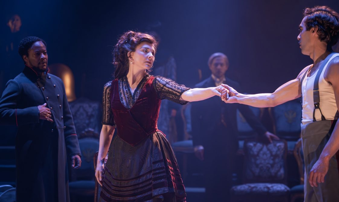 A group of performers gathered. The man on the right is holding hands with the woman in the centre. They are looking into each other's eyes deeply. There is a man on the left who is looking at them with a pained expression; his hand is resting on his stomach. There are several other characters in the background looking at the three people at the front of the scene.